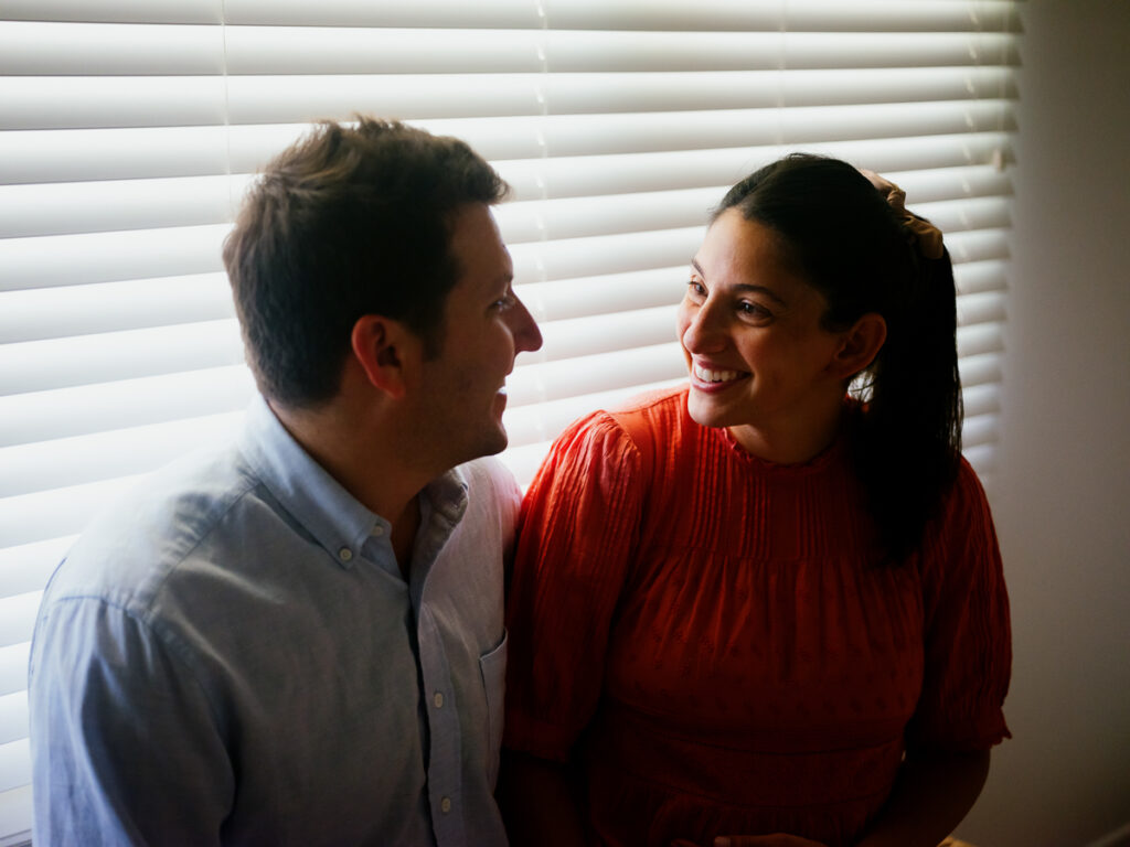 A young couple looks at each other and smiles in a dimly lit room, she wears a bright orange dress, he wears a blue button-up shirt. 