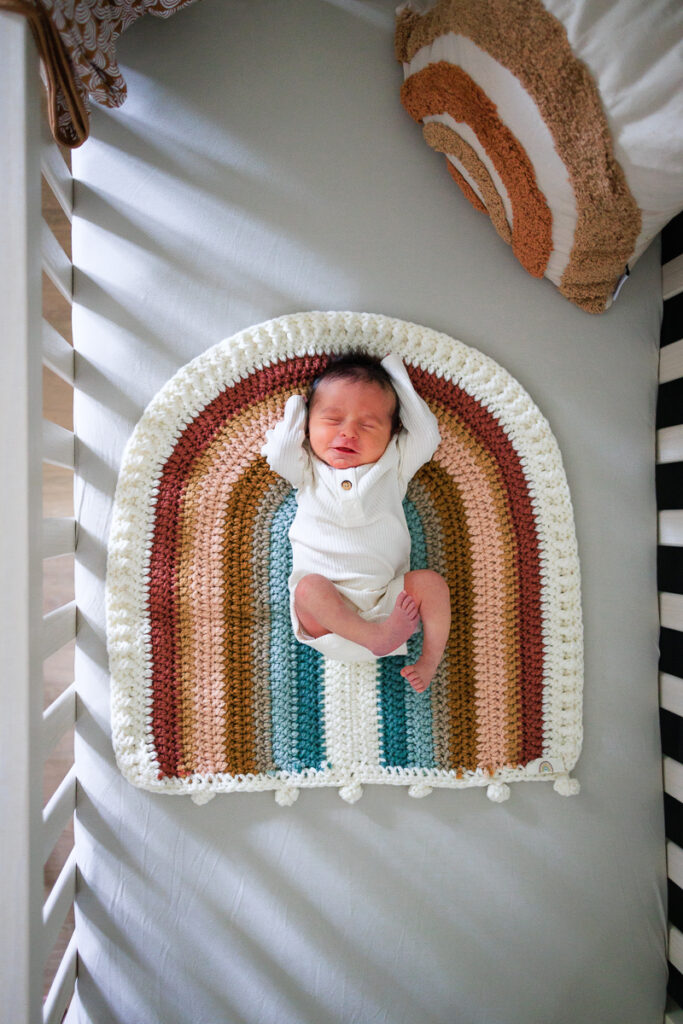 Newborn baby lays in his crib over a crochet rainbow blanket with muted colors, a matching pillow in the corner. 