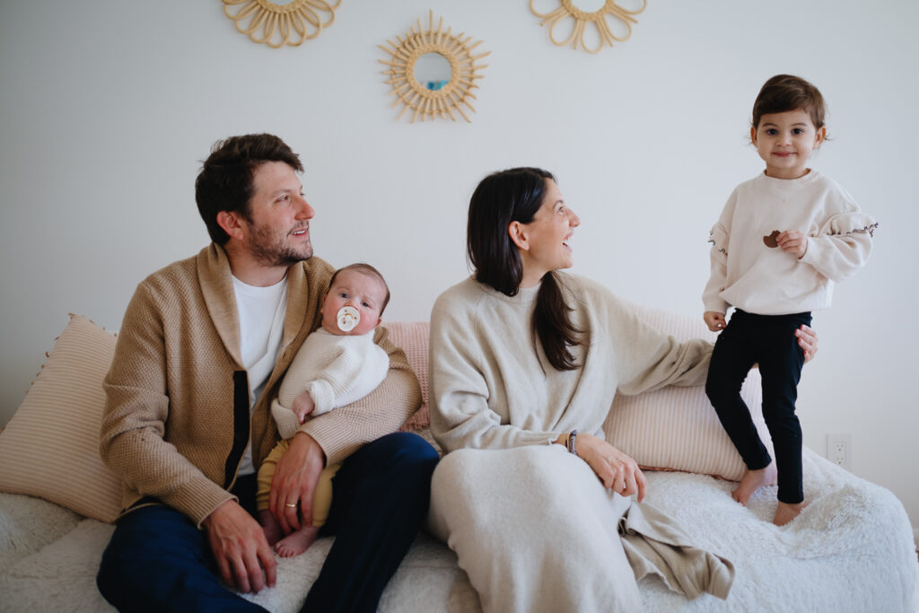 Family of four together at home, all wearing neutral tones that complement each other providing the perfect example of what to wear for a newborn photo session. 