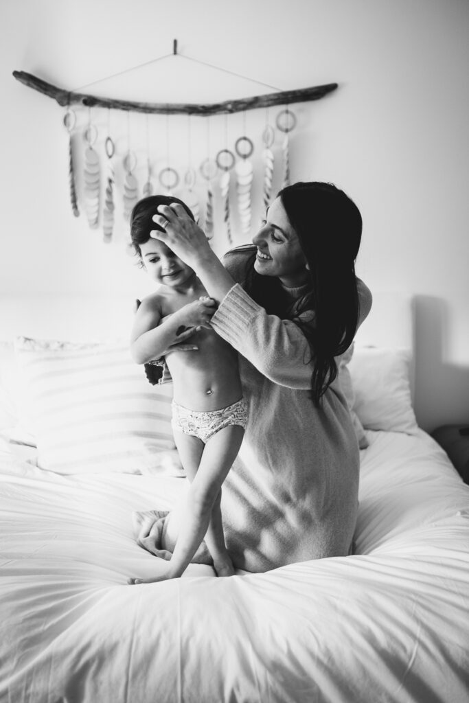 Black & white image of a mother playfully changing her daughter's outfit on her bed. 