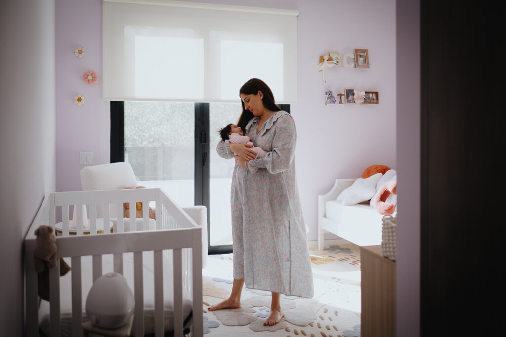 A new mom rocking her newborn baby in a pink & purple nursery. Mom wears a moo moo with equally soft and pastel colors, complementing the palette of the room.