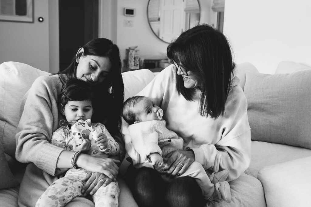 Black & white image of a young mom with her infant and toddler and her mother all cuddled together on the couch. 