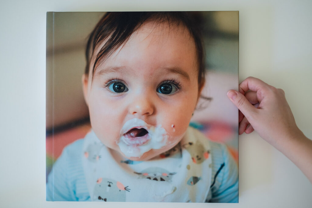 Overhead picture of a photo album with an image cover of a 6-month-old baby with yogurt all over her face. A hand reaches over from the side as it to flip through the album. 