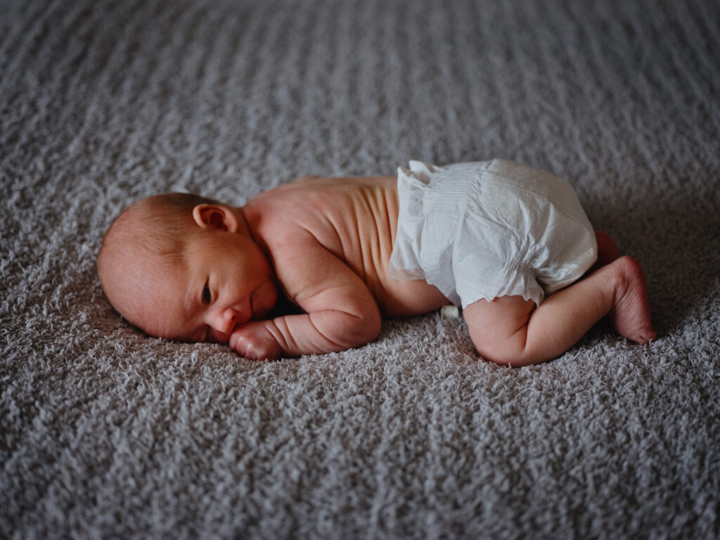 Newborn baby down to the diaper lying on a grey, textured blanket. 