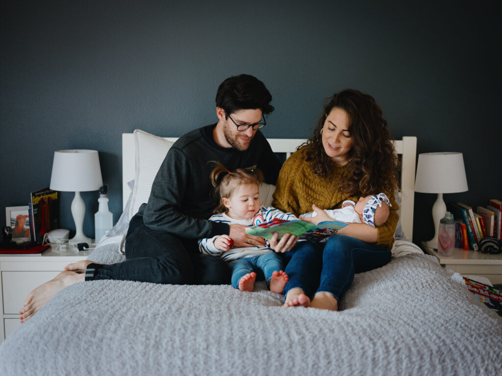 Family of 4 sit on the bed - mom, dad, a toddler, and newborn baby. They read a book together.