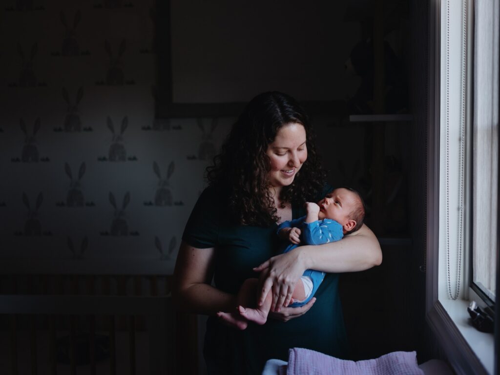 Self portrait of photographer Marjorie Cohen holding her newborn baby. They are in a pocket of light in an otherwise dimly lit room.