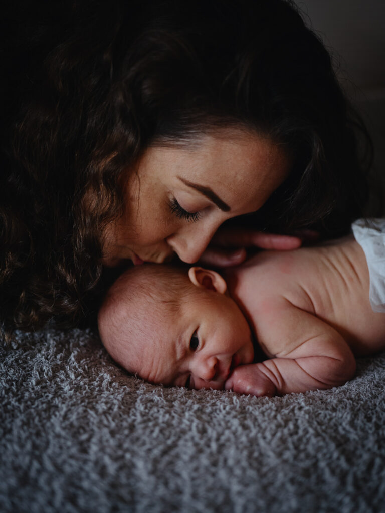 A mother kisses the head of her newborn baby, who lays on a textures grey blanket.