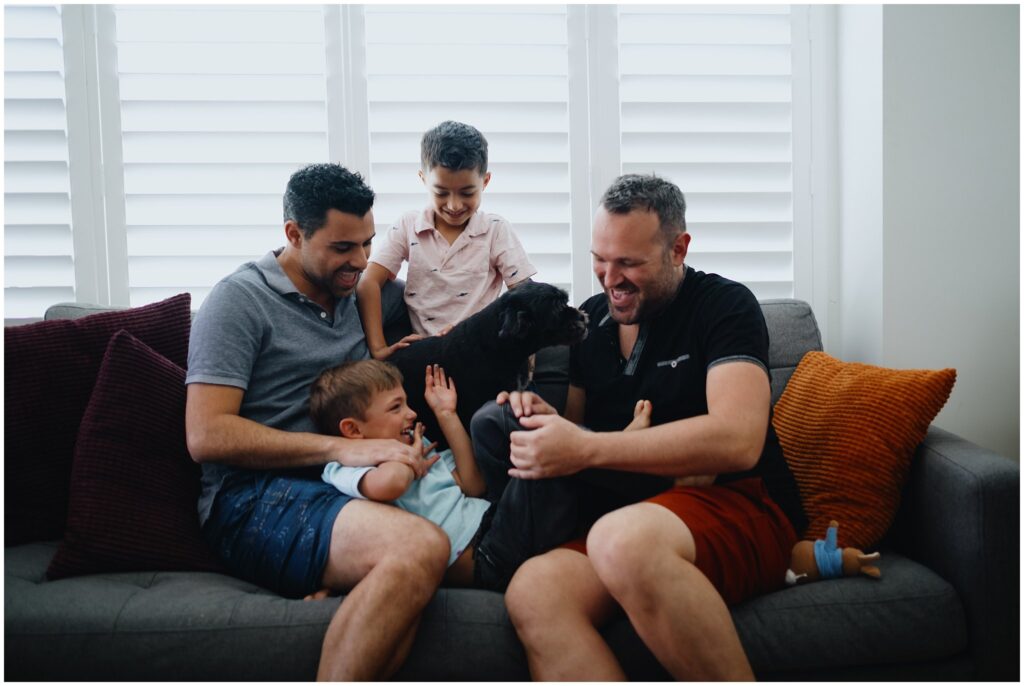 A family of four, two dads and their two 5-year-old twin sons, plus their black pup, playfully snuggling on the couch. An example of how everyday clothes can still work for your family photography session. 