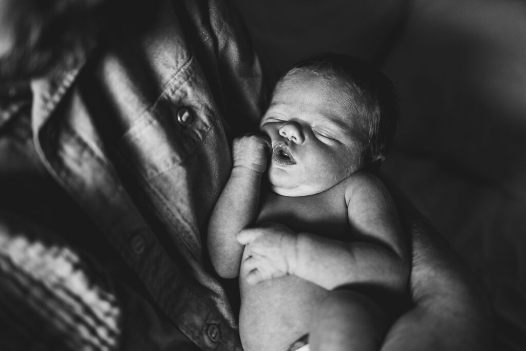 Black & white photo of a newborn baby in his parent's lap, his little hand pushing up his cheek.