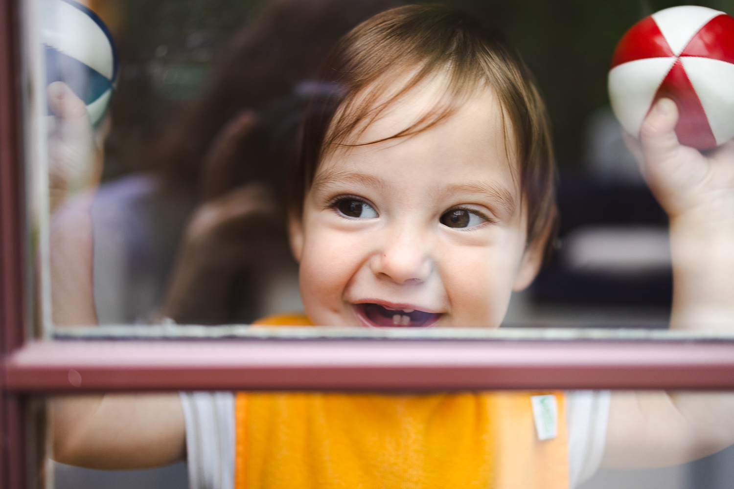 Photo taken through a window of a baby looking out and smiling big, his two bottom teeth showing.