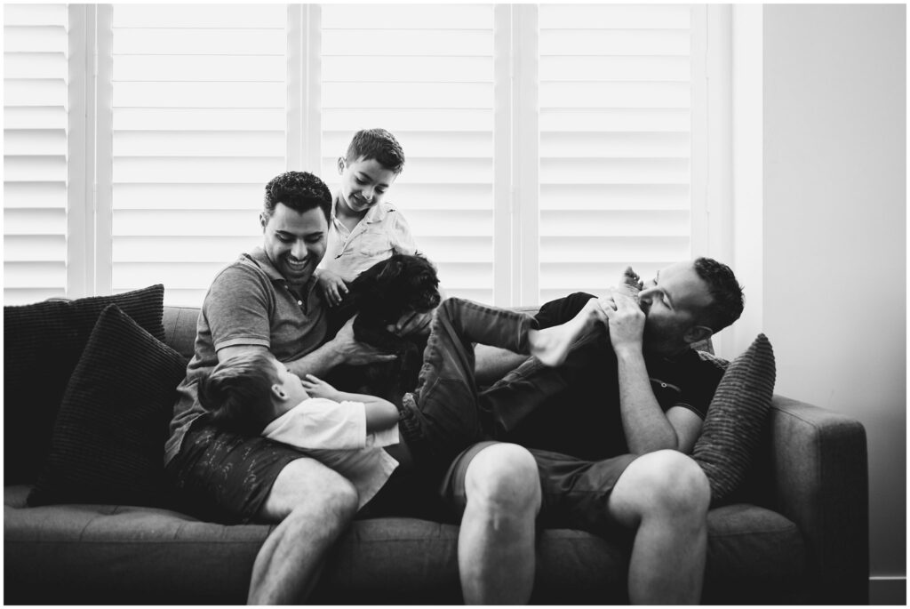 Black & white photo of a family of four interacting playfully on their couch: two dads and two twin boys. 