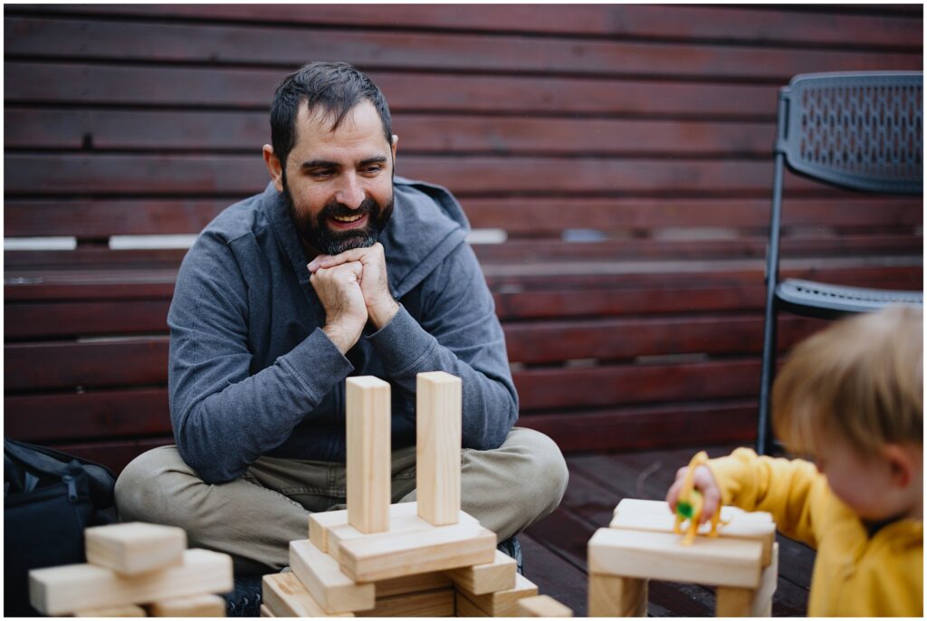 A dad happily watching his toddler son playing with blocks on their outside patio. 
