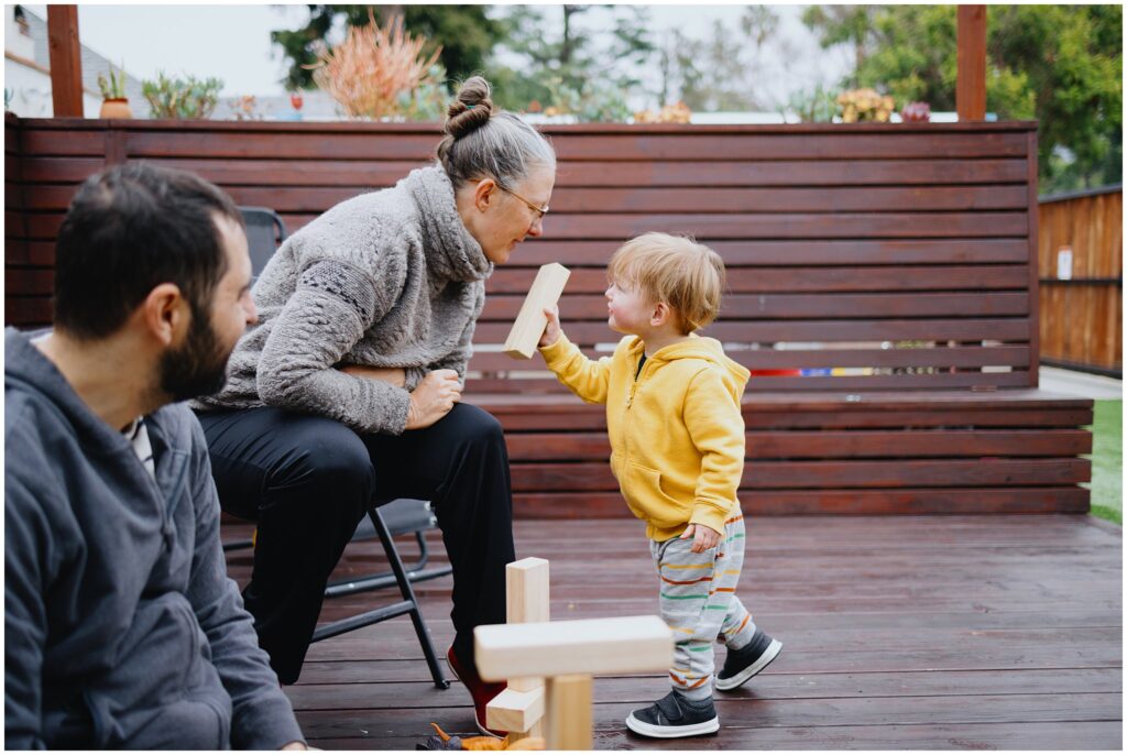 A toddler boy in a yellow jacket showing his mom one of his wooden blocks on their outdoor patio. Dad watches from the side. 