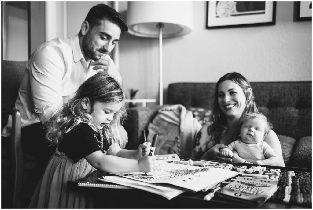 Black & white photo of family of 4 - mom, dad, toddler girl, and newborn baby boy, coloring together in their living room. 