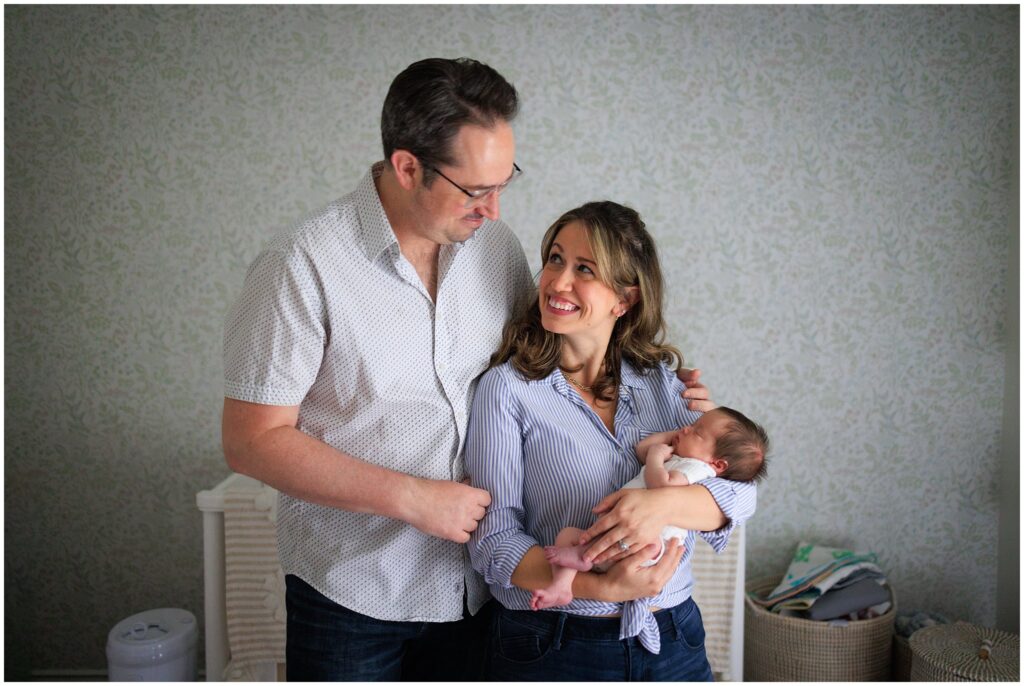 Newborn parents hold their baby in the nursery in front of a floral wallpaper. 