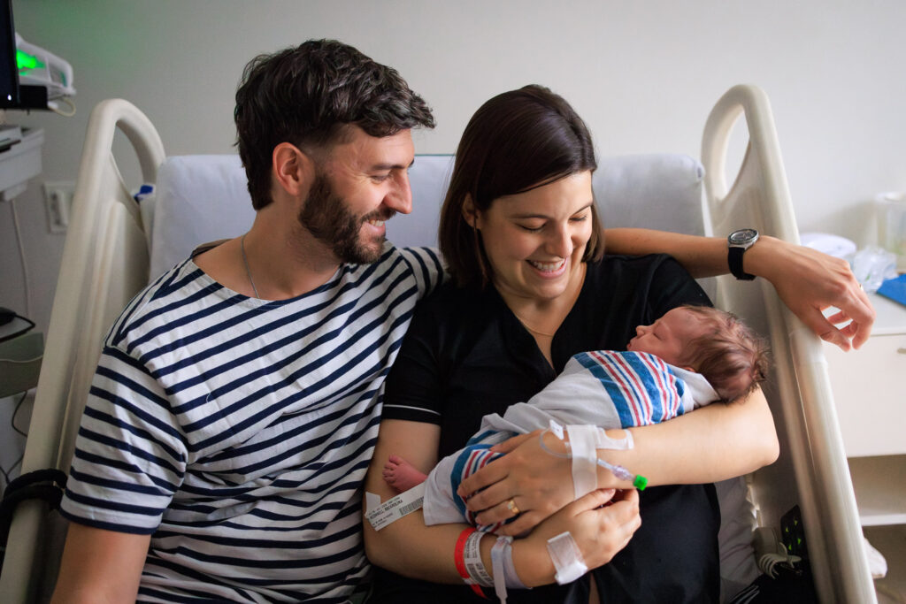 Parents giddily gaze down at their newborn baby, all squeezed together in mom's hospital bed.